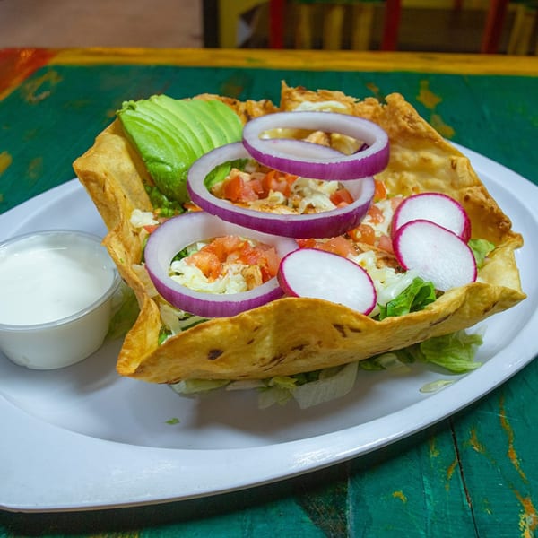 Taco salad in a crispy tortilla bowl with avocado, red onion, radishes, and a side of sour cream.