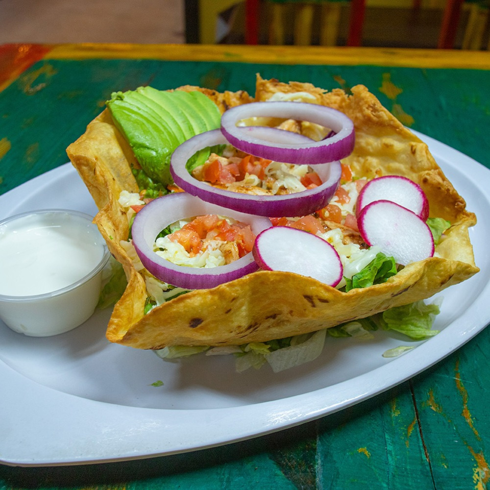 Taco salad in a crispy tortilla bowl with avocado, red onion, radishes, and a side of sour cream.