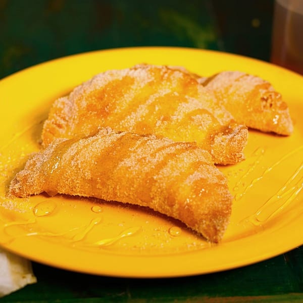 Three golden-brown, sugar-dusted empanadas drizzled with honey on a bright yellow plate.