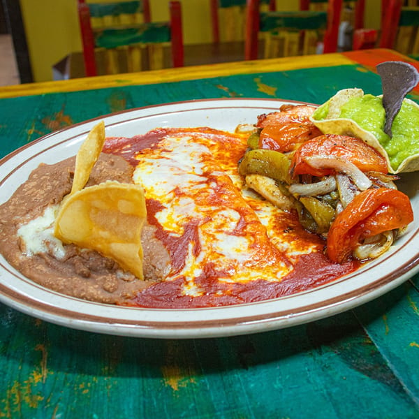 Mexican dish with refried beans, cheesy enchiladas, fajita vegetables, and guacamole on a colorful table.