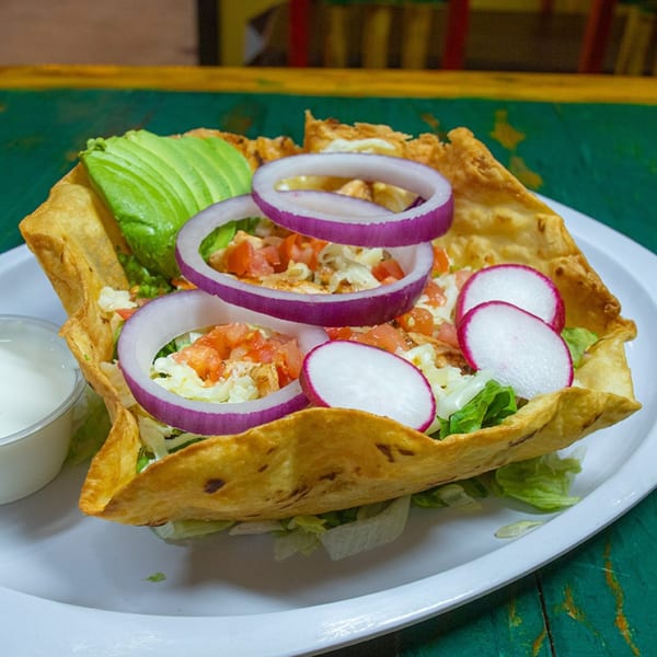 Taco salad in a crispy shell with avocado, red onion, radishes, and a side of dressing.