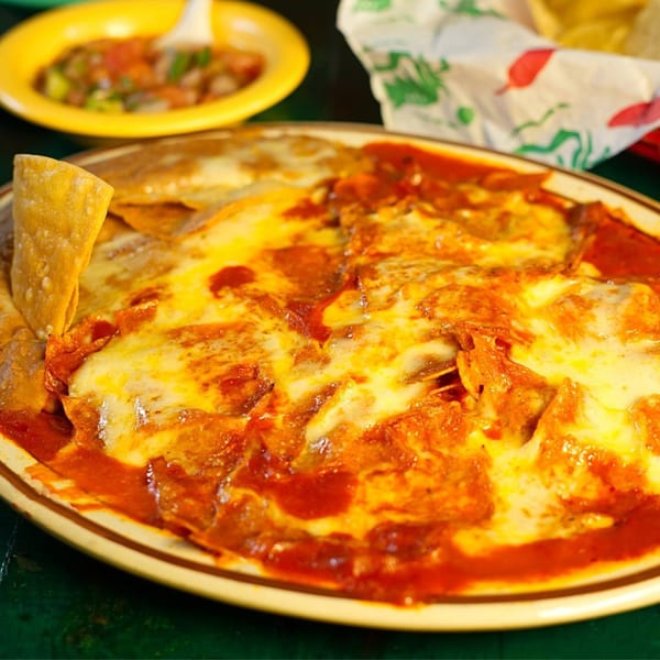 A plate of cheesy enchiladas with red sauce, a tortilla chip, and salsa in the background.