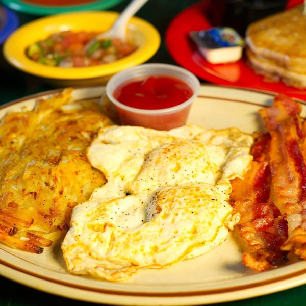 Breakfast plate with hash browns, two fried eggs, bacon, and a side of ketchup.